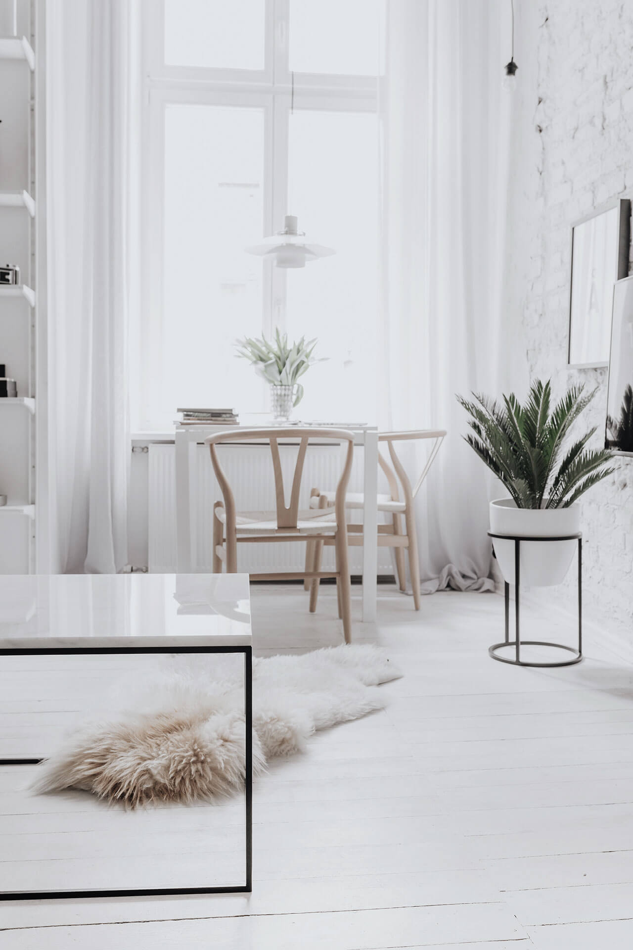 Bright minimalist interior with white walls and floors, a large window with sheer curtains, a small dining table with two wooden chairs and a potted plant, a larger plant on a stand, a fluffy rug, a reflective coffee table, and shelves with various items.