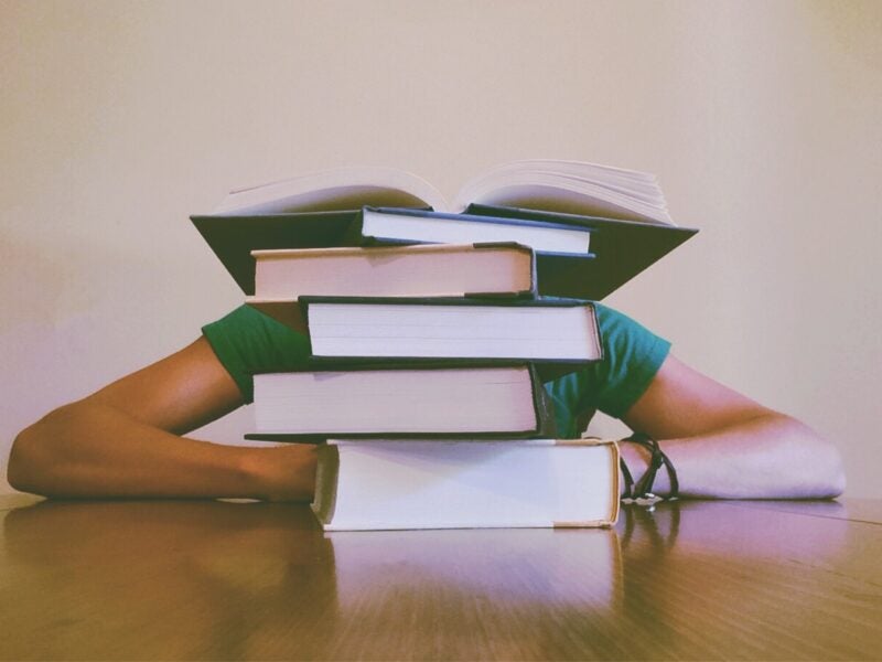 Image of someone leaning against a table with their head down, obscured by a pile of four closed books, with an open book at the top