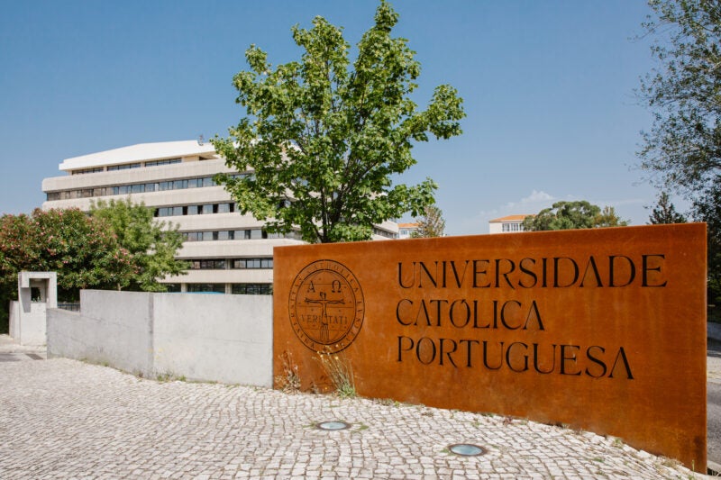 The image shows the entrance to Universidade Católica Portuguesa, with a large sign displaying the university's name and emblem. The background includes a modern building and trees.