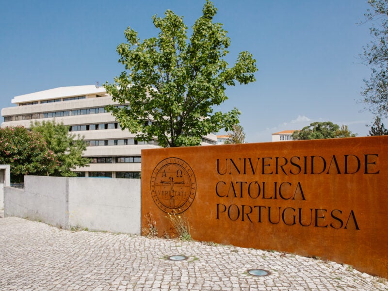 The image shows the entrance to Universidade Católica Portuguesa, with a large sign displaying the university's name and emblem. The background includes a modern building and trees.
