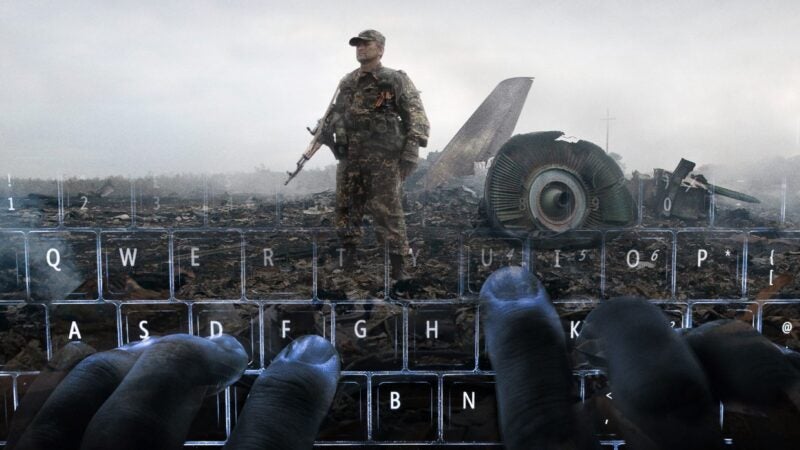 The image shows a soldier standing in a wreckage site with debris and parts of an aircraft visible in the background. The foreground features hands typing on a keyboard, creating an overlay effect with the scene behind it.