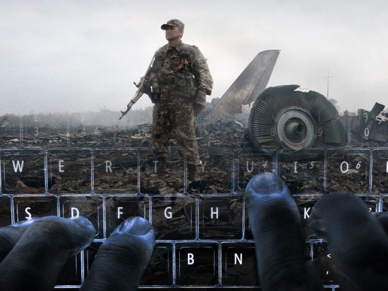 The image shows a soldier standing in a wreckage site with debris and parts of an aircraft visible in the background. The foreground features hands typing on a keyboard, creating an overlay effect with the scene behind it.