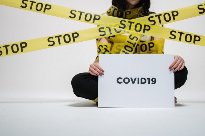 The image shows a person sitting cross-legged on the floor, holding a sign that reads "COVID19." The person is wrapped in yellow caution tape with the word "STOP" written on it multiple times.