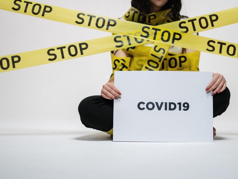 The image shows a person sitting cross-legged on the floor, holding a sign that reads "COVID19." The person is wrapped in yellow caution tape with the word "STOP" written on it multiple times.