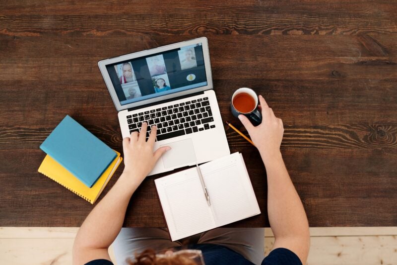 The image shows a person sitting at a wooden table, using a laptop for a video call. The laptop screen displays four participants in the video call. The person has an open notebook with a pen placed diagonally on it, and is holding a cup of tea or coffee in the other hand. There are also two closed notebooks, one blue and one yellow, placed on the table next to the laptop.
