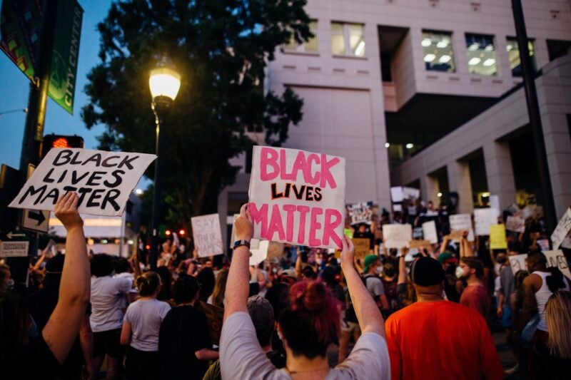 The image shows a large group of people gathered in a protest. Many individuals are holding signs, with the most prominent ones reading "BLACK LIVES MATTER." The protest appears to be taking place in an urban area, possibly near a government or office building, as indicated by the architecture and windows in the background. There is also a street lamp illuminating part of the scene.
