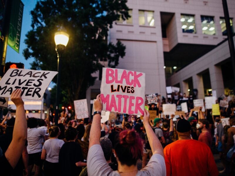 The image shows a large group of people gathered in a protest. Many individuals are holding signs, with the most prominent ones reading "BLACK LIVES MATTER." The protest appears to be taking place in an urban area, possibly near a government or office building, as indicated by the architecture and windows in the background. There is also a street lamp illuminating part of the scene.