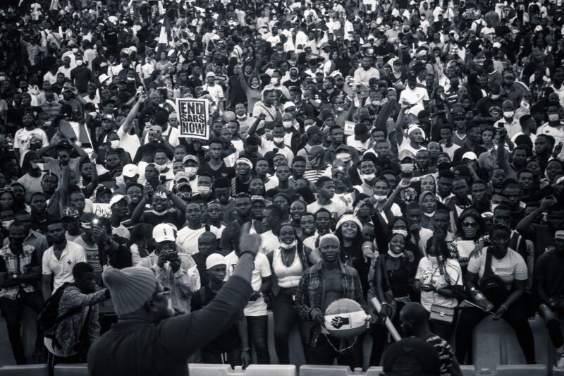 The image shows a large crowd of people, many of whom are holding up their phones. One person in the foreground is holding a sign that reads "END SARS NOW." The crowd appears to be participating in a protest or rally.