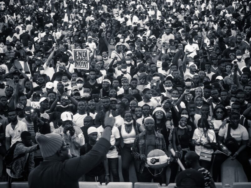 The image shows a large crowd of people, many of whom are holding up their phones. One person in the foreground is holding a sign that reads "END SARS NOW." The crowd appears to be participating in a protest or rally.