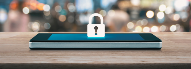 The image shows a smartphone lying flat on a wooden surface with a digital lock icon displayed prominently on the screen. The background is blurred.