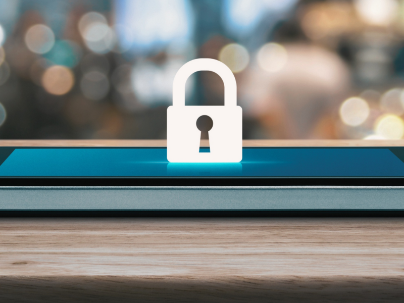 The image shows a smartphone lying flat on a wooden surface with a digital lock icon displayed prominently on the screen. The background is blurred.