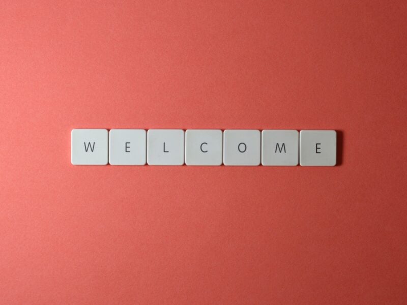 The image shows seven white square tiles arranged in a horizontal line on a red background. Each tile has a single black letter, collectively spelling out the word "WELCOME".