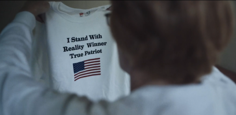 The image shows a person holding up a white t-shirt. The t-shirt has text and an image of the American flag printed on it. The text reads: "I Stand With Reality Winner True Patriot".