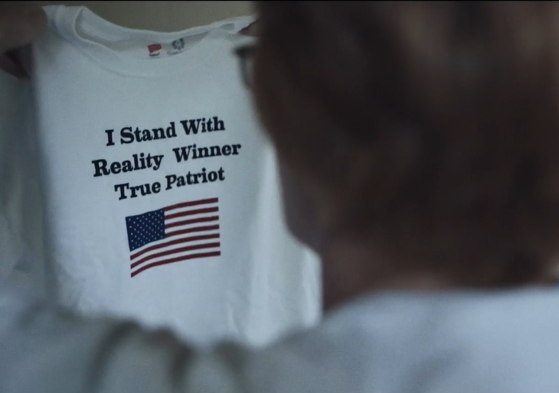 The image shows a person holding up a white t-shirt. The t-shirt has text and an image of the American flag printed on it. The text reads: "I Stand With Reality Winner True Patriot".