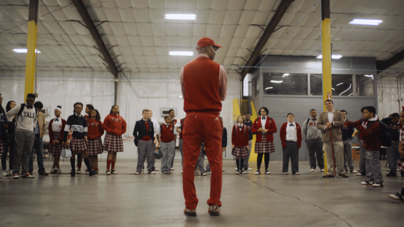 A group of students and adults standing in a semi-circle inside a large indoor space with high ceilings. The central figure, seen from behind, wears red clothing including a cap, vest, and pants. The students are in school uniforms with plaid skirts or pants and red sweaters or blazers