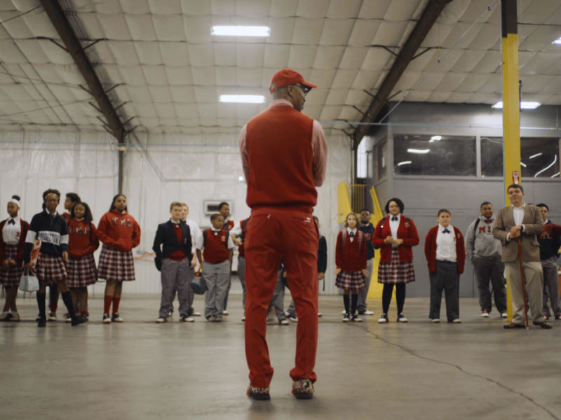 A group of students and adults standing in a semi-circle inside a large indoor space with high ceilings. The central figure, seen from behind, wears red clothing including a cap, vest, and pants. The students are in school uniforms with plaid skirts or pants and red sweaters or blazers