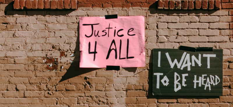 Two protest signs taped to a brick wall. The left sign is pink with black text reading 'Justice 4 ALL,' and the right sign is dark green with white text reading 'I WANT TO BE HEARD