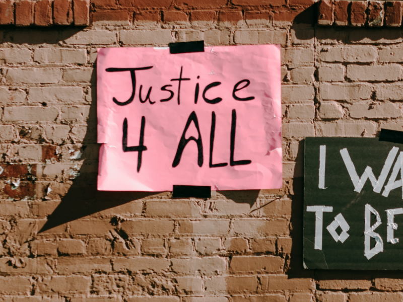 Two protest signs taped to a brick wall. The left sign is pink with black text reading 'Justice 4 ALL,' and the right sign is dark green with white text reading 'I WANT TO BE HEARD