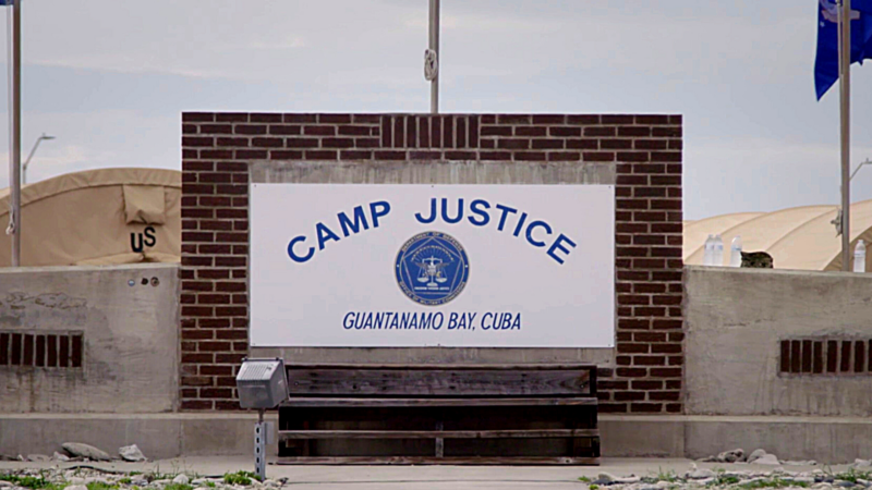 The image shows a sign that reads "CAMP JUSTICE GUANTANAMO BAY, CUBA" with a seal in the center. The sign is mounted on a brick wall, and there are tents and other structures visible in the background.