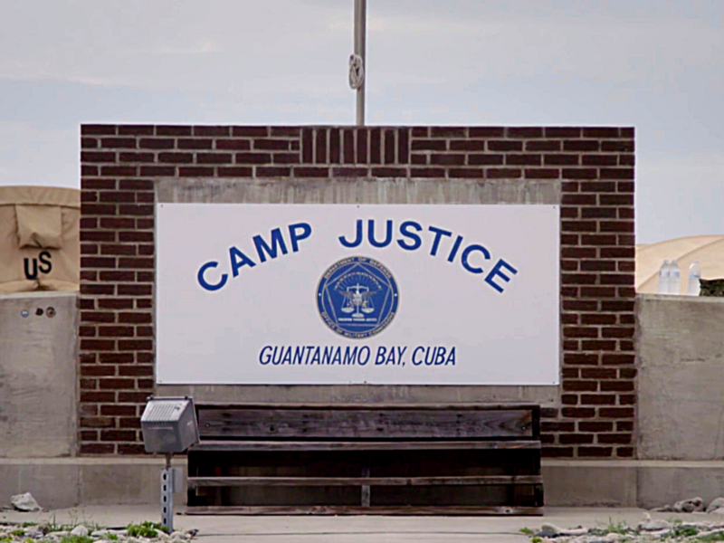 The image shows a sign that reads "CAMP JUSTICE GUANTANAMO BAY, CUBA" with a seal in the center. The sign is mounted on a brick wall, and there are tents and other structures visible in the background.