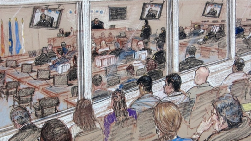 The image shows a courtroom scene with several people seated and observing the proceedings. There are multiple monitors displaying the judge and other individuals involved in the trial. The room is divided by a glass partition, separating the observers from those directly participating in the trial. There are flags visible on one side of the room, and various documents and laptops are placed on desks within the courtroom.