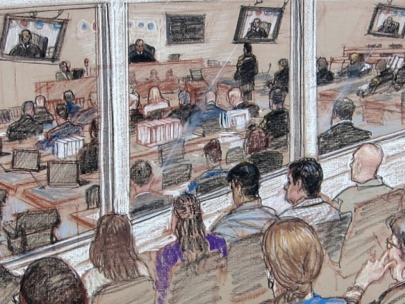 The image shows a courtroom scene with several people seated and observing the proceedings. There are multiple monitors displaying the judge and other individuals involved in the trial. The room is divided by a glass partition, separating the observers from those directly participating in the trial. There are flags visible on one side of the room, and various documents and laptops are placed on desks within the courtroom.