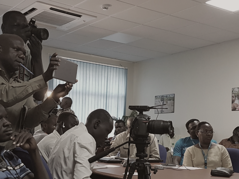 The image shows a group of people in a room, some of whom are taking photographs or recording videos. There are several cameras and recording devices visible.The room has fluorescent lighting and there is a window with vertical blinds in the background. There are also some posters on the walls.