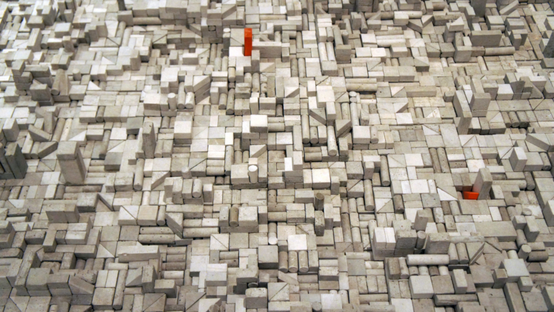 The image shows a complex, abstract arrangement of small, rectangular blocks and shapes in various shades of gray. There are also a few small red blocks scattered throughout the composition. The overall appearance resembles a densely packed, three-dimensional mosaic or cityscape.