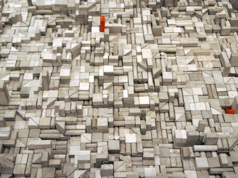 The image shows a complex, abstract arrangement of small, rectangular blocks and shapes in various shades of gray. There are also a few small red blocks scattered throughout the composition. The overall appearance resembles a densely packed, three-dimensional mosaic or cityscape.