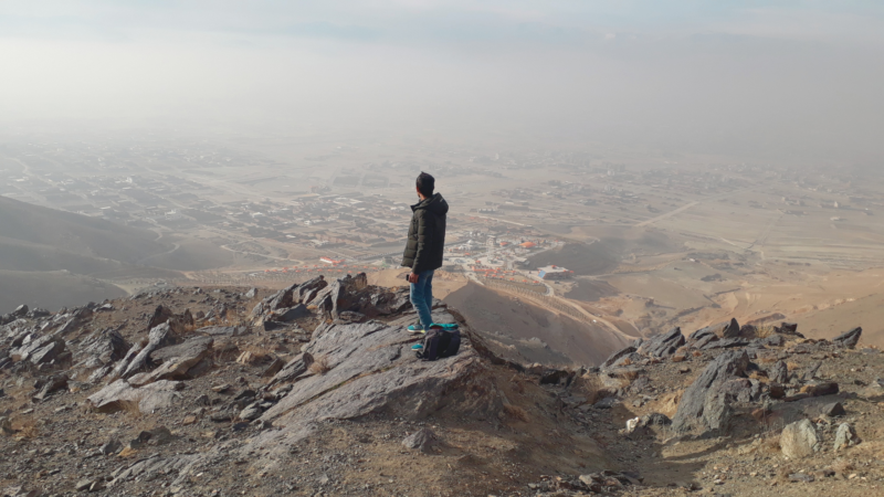 The image shows a person standing on a rocky terrain at an elevated viewpoint, overlooking a vast landscape that includes a city or town in the distance. The sky appears hazy, and the person has a backpack placed on the ground beside them.