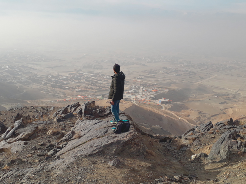 The image shows a person standing on a rocky terrain at an elevated viewpoint, overlooking a vast landscape that includes a city or town in the distance. The sky appears hazy, and the person has a backpack placed on the ground beside them.
