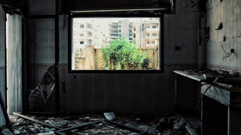 The image shows an abandoned room with a broken window and door. The room is filled with debris, including broken glass, papers, and other materials scattered on the floor. Outside the window, there is a view of buildings and greenery.