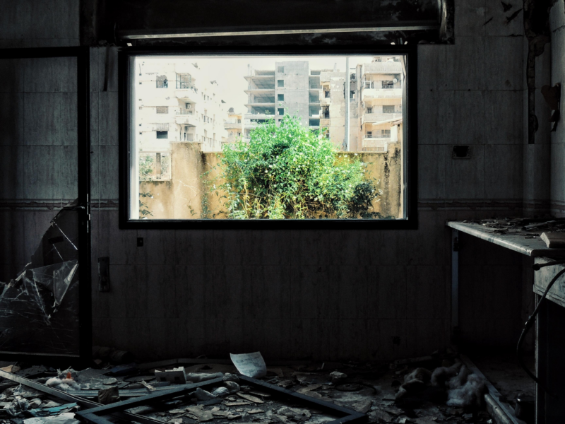 The image shows an abandoned room with a broken window and door. The room is filled with debris, including broken glass, papers, and other materials scattered on the floor. Outside the window, there is a view of buildings and greenery.
