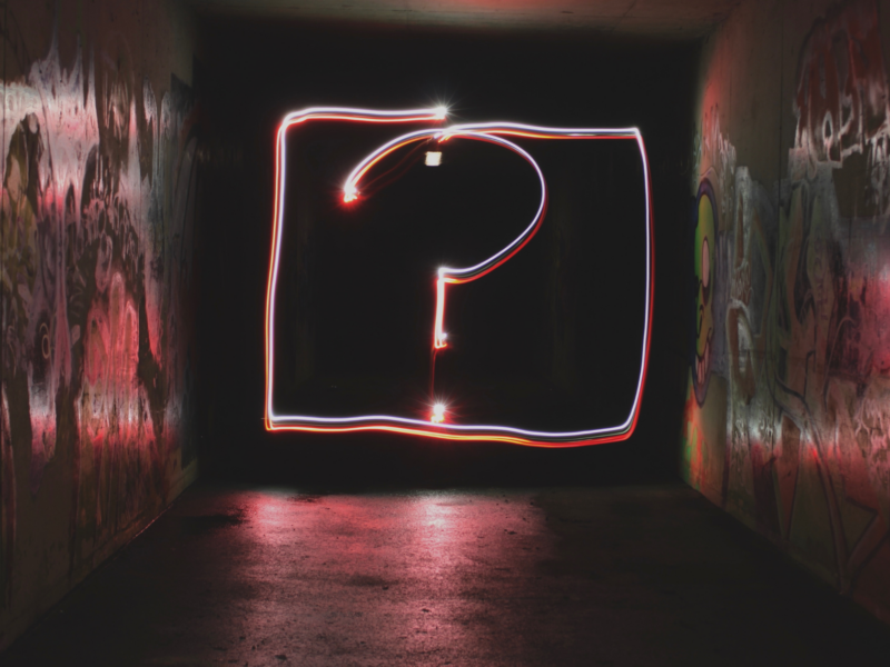 The image shows a dark tunnel with graffiti on the walls. In the center of the tunnel, there's a large question mark outlined with light, creating a glowing effect. The question mark is enclosed within a rectangular frame also made of light.