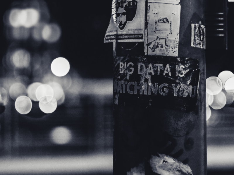 The image shows a black and white photograph of a pole with various posters attached to it. The most prominent poster reads "BIG DATA IS WATCHING YOU." The background is blurred.