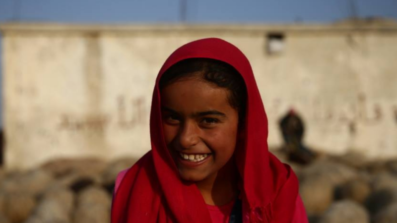 The image shows a person wearing a red headscarf and a pink shirt standing in front of a wall with some text on it.