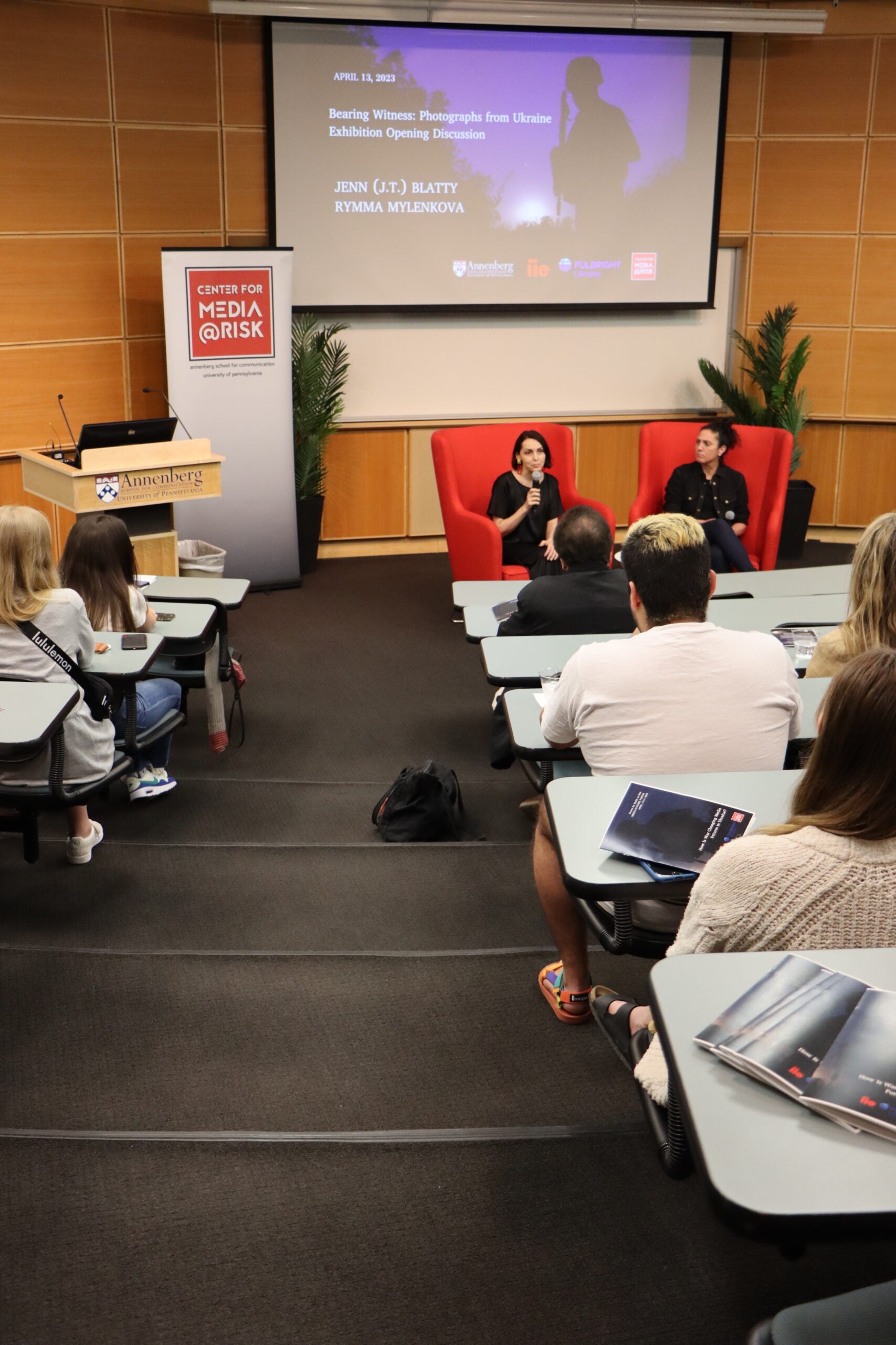the image shows a classroom or lecture hall setting with several people seated at desks, facing the front of the room. There is a large screen displaying an image of people in conversation, and two individuals are seated at the front, possibly leading a discussion or presentation. The room has modern lighting and design elements, including wooden panels on one wall and acoustic panels on another.