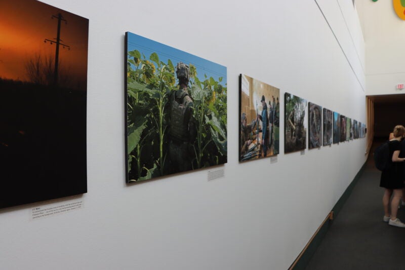 The image shows a hallway with a series of photographs displayed on the wall. The photographs appear to be part of an exhibition, each mounted on the wall in a row. There are two people visible at the end of the hallway, one of whom is wearing a backpack. The lighting in the hallway is bright, and there is text beneath each photograph, likely descriptions or titles for each piece.