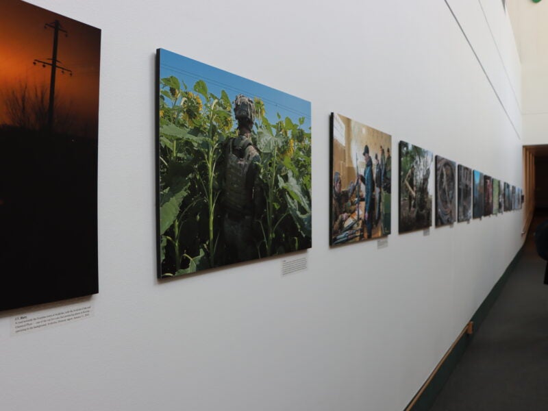 The image shows a hallway with a series of photographs displayed on the wall. The photographs appear to be part of an exhibition, each mounted on the wall in a row. There are two people visible at the end of the hallway, one of whom is wearing a backpack. The lighting in the hallway is bright, and there is text beneath each photograph, likely descriptions or titles for each piece.