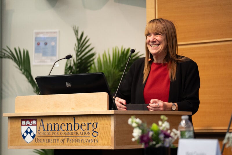 Barbie Zelizer, wearing red, and smiling from the lectern at Annenberg.