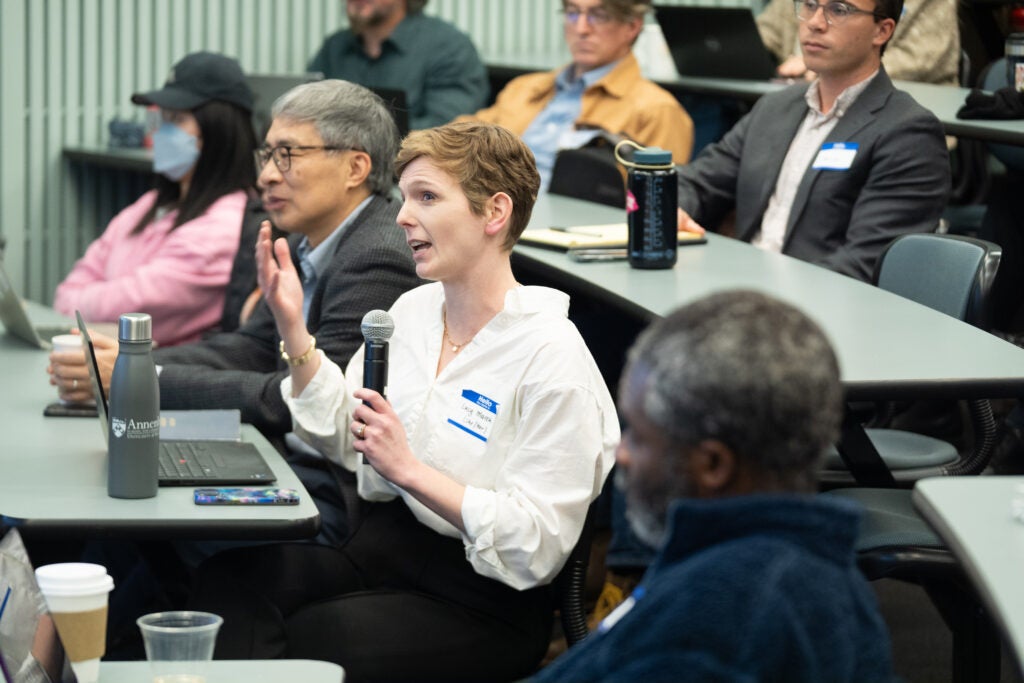 An audience member wearing a white shirt asking a question during a conference, holding a microphone and gesturing.