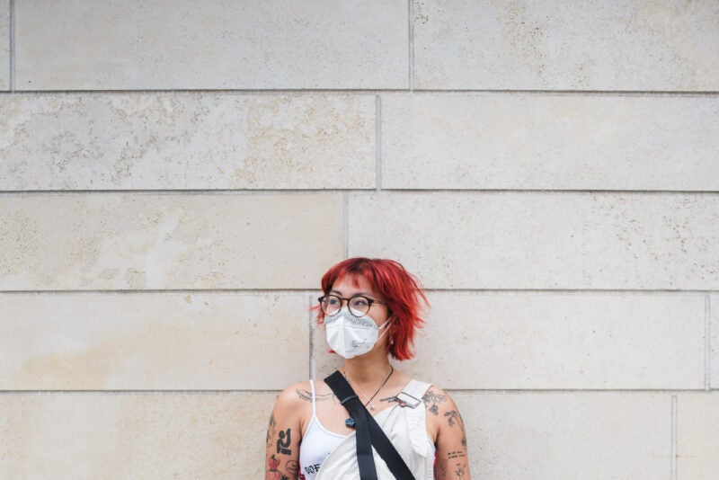 Image of James Salanga standing in front of a white wooden wall with red hair, glasses, and wearing a white mask. They are wearing a white top with a black strap visible, perhaps from a cross body bag. They are looking away from the camera at an angle. Photo by Abby Mahler.