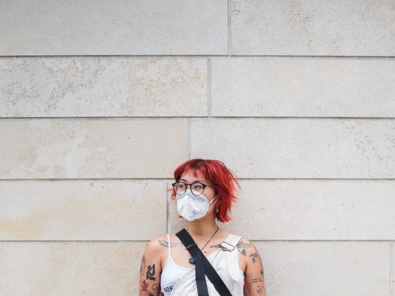 Image of James Salanga standing in front of a white wooden wall with red hair, glasses, and wearing a white mask. They are wearing a white top with a black strap visible, perhaps from a cross body bag. They are looking away from the camera at an angle. Photo by Abby Mahler.