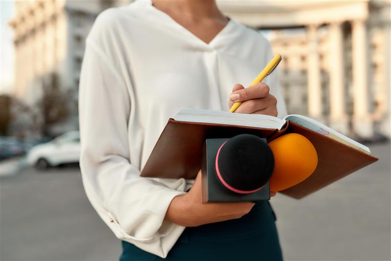 A person holding an open notebook and writing with a yellow pen, while also carrying two handheld microphones, standing outdoors in an urban setting with blurred buildings in the background.
